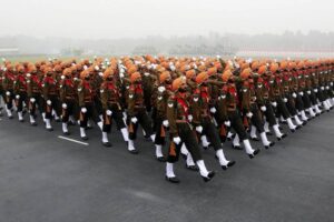 A large group of Sikh soldiers in orange turbans and khaki uniforms march in perfect formation on a wide road during a parade, moving in unison with disciplined posture against a foggy backdrop.