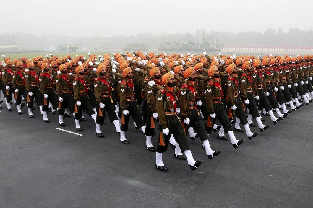 A large group of Sikh soldiers in orange turbans and khaki uniforms march in perfect formation on a wide road during a parade, moving in unison with disciplined posture against a foggy backdrop.